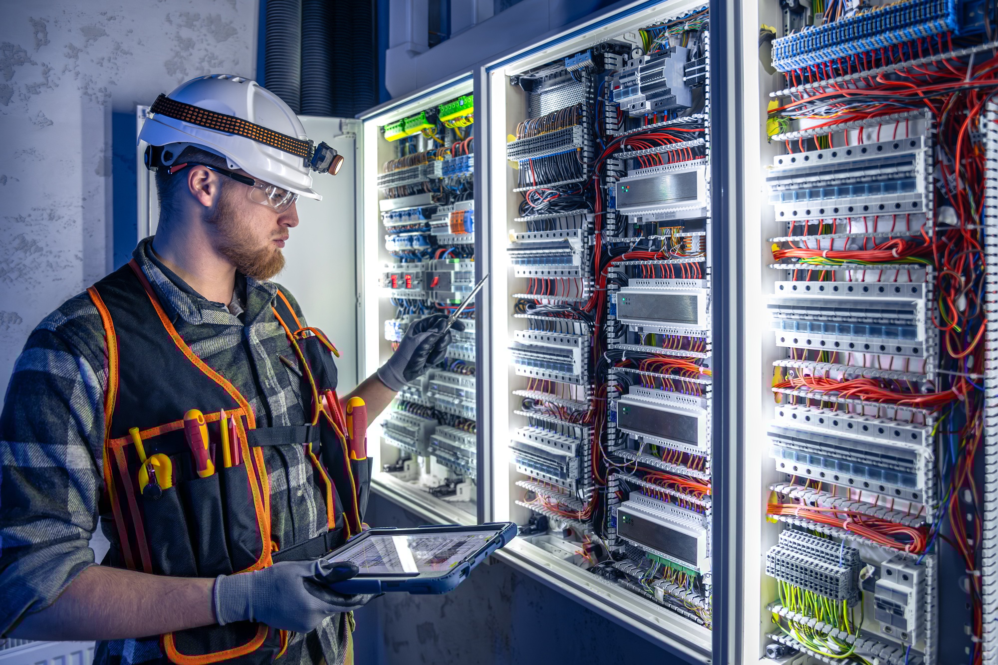 Electrician in overalls, focused on work in switchboard with fuses, using tablet.